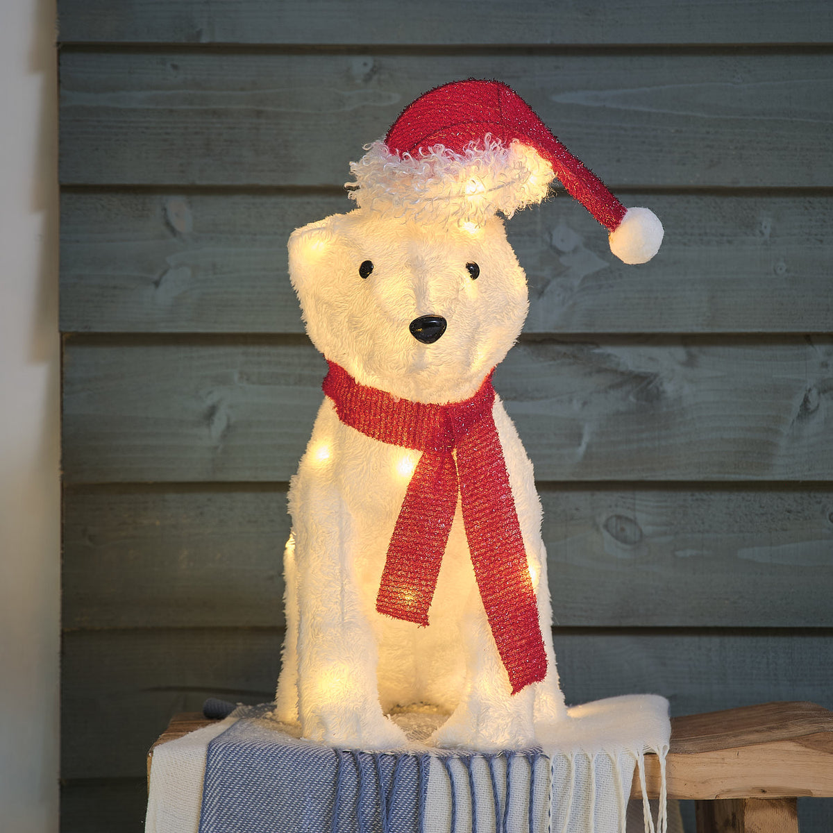 Un ours en peluche illuminé portant un bonnet rouge et une écharpe rouge, assis sur un socle gris devant un fond de mur en bois gris.